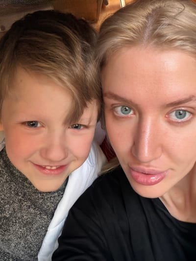 The image captures a close-up portrait of an adult female and a young male child, located in Goričani, Montenegro. The female, positioned on the right, has fair skin, blonde hair, and blue eyes, with full lips and a neutral to slightly smiling expression. She is wearing a black top. The male child, positioned on the left, has fair skin, light brown hair, and blue eyes. He displays a broad smile, showing his teeth, and is wearing a grey textured top with a white collar visible beneath. Both individuals are looking directly into the camera. The background is blurred and indistinct, suggesting an indoor setting with hints of light brown and reddish-brown surfaces.