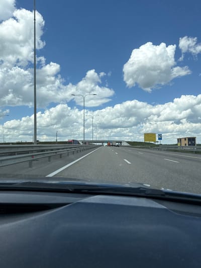 The image shows a view from inside a vehicle, likely a car, driving along a highway. The highway appears to be multi-lane and asphalt. The road is lined with streetlights. A guardrail is visible. In the distance, there are signs posted and a building. The sky is predominantly blue with numerous white, fluffy clouds. The exact location is Grushevskaya, Russia. There is no indication of human presence.