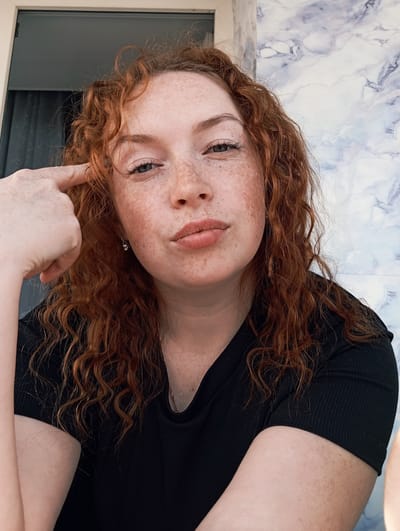 A close-up, rotated portrait shows a young person with vibrant, curly red hair and numerous freckles scattered across their face. The person is lying down, looking upwards towards the camera with a subtle, contemplative expression. Their head is tilted, and one hand is raised, with a finger gently touching the hair or ear area.
They are wearing a ribbed black top, visible around the shoulders and neck. A delicate necklace with a small pendant is also visible on their chest. Their eyes are a light color, possibly blue or grey, and their lips are slightly parted.
The background primarily features a white and blue marbled pattern, which appears to be a wall or ceiling surface, indicating an indoor setting. Below the person's head, a darker, flat surface, possibly a window frame or dark furniture, is partially visible. The lighting is soft and even, suggesting it is not direct sunlight. There is no visible text in the image. The overall mood is calm and introspective.