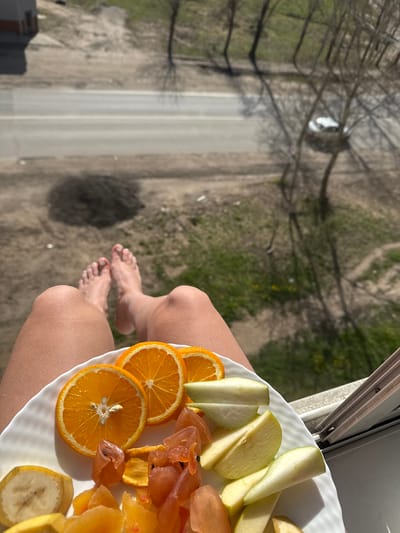 The image, set in Novo-Nikolskoye, Russia, captures a high-angle perspective from above, looking down onto a street and the surrounding landscape. In the immediate foreground, the lower half of a person's body is visible, featuring bare legs and feet, suggesting they are seated or reclining on a balcony or by an open window.
The person holds a white plate, which is prominently displayed and filled with a variety of fresh and dried fruits. Visible on the plate are several bright orange slices, pieces of green apple, slices of banana, and a collection of translucent, amber-toned dried fruits, possibly apricots or persimmons.
The background reveals a street with two distinct lanes, partially obscured by the bare branches of dormant trees. The ground adjacent to the road is unpaved, showing patches of dry earth and some green grass. A small, light-colored car is faintly visible in the distance on the road, partially hidden by the tree canopy. A dark, circular shadow, possibly cast by the person's head or hair, is visible on the ground below.
The scene is bathed in bright natural sunlight, indicating daytime, likely morning or afternoon, with clear weather. The bare trees suggest either early spring or late autumn. There is no visible text in the image. The overall impression is one of a leisurely moment, enjoying a fruit snack outdoors with an expansive view.