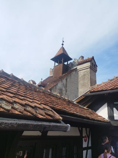 This media file captures an exterior view of historic architecture with a bright, clear sky overhead, suggesting fair weather and daytime. The scene is dominated by terracotta tiled roofs, some appearing weathered and moss-covered, indicative of age. In the background, a stone tower with a wooden structure at its peak rises above the rooftops, topped with a weather vane. Adjacent to it is a tall, square stone chimney with a layered top, also capped with tiles. The architecture suggests a European historical setting, possibly a castle or an old village.
In the foreground, the eaves of a building with dark wooden beams and white infill are visible, hinting at traditional construction. The lower portion of the image shows glimpses of people, with the back of a woman in a purple top and a man with glasses visible. They appear to be observing something, though their expressions are not discernible. A red and white circular sign is partially visible on the building's facade.
The overall impression is one of exploring a historical site. The clarity of the sky and the brightness of the light suggest it is a sunny day, likely late morning or early afternoon. The location context of Predeluț, Romania, further points to a site of historical and possibly touristic interest within the region. No discernible text is visible in the image beyond the small circular sign, which is not legible.