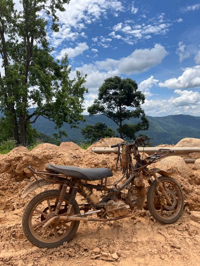 An abandoned, rusted motorcycle is parked on a dirt mound. The motorcycle is covered in dirt and debris, with its seat and handlebars visible. In the background, lush green trees and rolling hills create a scenic backdrop, under a bright blue sky with fluffy white clouds. The scene is set in a rural area, likely in the vicinity of Nao Se, Vietnam, given the landscape and vegetation. There are no people present in the image. The motorcycle appears to be a dirt bike or similar off-road model, its condition suggesting it has been exposed to the elements for an extended period. The surrounding terrain is rugged and uneven, composed primarily of reddish-brown soil and rocks, indicative of a construction site or a natural landslide area.