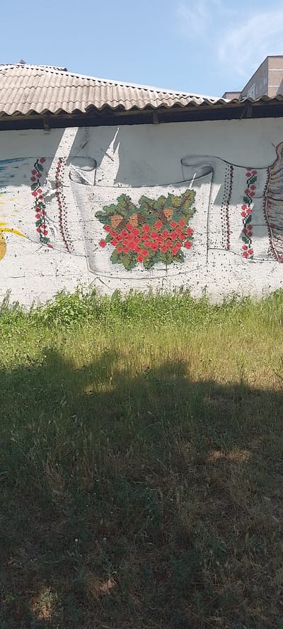 The image shows a mural on the exterior wall of a building, depicting a floral motif and traditional Ukrainian embroidery patterns. The mural is rendered in a folk art style, with vibrant red and green colors used for the flowers and intricate geometric designs for the borders. The wall itself appears to be made of concrete or plaster and is weathered, with some visible cracks and chips. Above the wall is a corrugated metal roof, characteristic of many industrial or utilitarian buildings. The foreground is dominated by tall, overgrown grass, suggesting a neglected or semi-rural setting. The sky is clear and blue, indicating a sunny day. The architectural style of the adjacent building on the right suggests an urban environment. The overall impression is one of a public space decorated with local cultural elements. Based on the visual cues and the request to identify the location in Cherkasy, Ukraine, this mural is likely situated in an outdoor public area or on the side of a building within the city.