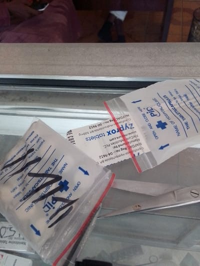 The image presents a close-up, downward-angled view of medical supplies arranged on a reflective surface, likely a countertop or display case. Three transparent plastic ziplock bags, designed for dispensing medication, are the primary focus.
The bag in the foreground, positioned towards the lower left, contains multiple white capsules. It features a red zipper closure and blue printed text, including "OPEN AND CLOSE HERE" with a directional arrow, a blue cross logo, "PID," and fields for "NAME OF HOSPITAL/CLINIC" and "THE TABLETS/CAPSULES." Black marker scribbles partially obscure some of this text. Another similar bag is partially visible above and to the right of the foreground bag.
A third bag, prominently displaying a white label, is positioned centrally. This label identifies its contents as "Zyprox tablets," specifying "Ciprofloxacin Hydrochloride B.P. 500mg." The label also details "Manufactured by: McCurre Industries PLC" and displays a "NAFDAC Reg no:- 04-9612," indicating the product is regulated by Nigeria's National Agency for Food and Drug Administration and Control.
To the right of the medication bags, a pair of silver-toned scissors with a gold-colored pivot screw is visible, partially obscured underneath the bags. The background above the counter features a beige, textured surface. Above this, a blurred reflection shows a person in a maroon or purple garment holding a white object, and a darker, indistinct shape, suggesting an indoor environment such as a pharmacy or clinic. The NAFDAC registration number confirms the location as Nigeria. The image contains no visual information to specifically identify the location within the city of Muhammadu Dikko Stadium.