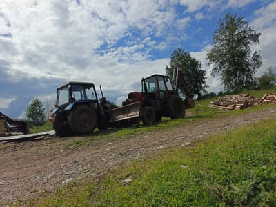 This media file depicts an outdoor scene with two tractors on a dirt road. The setting appears to be rural, possibly a farm or construction site, given the presence of stacked firewood in the background. The sky is cloudy with patches of blue, suggesting a partly sunny day.
The two tractors are the main subjects. The tractor on the left is blue and appears to be a utility tractor with a cab. It has a large bucket attachment in front. The tractor on the right is red and seems to be a backhoe loader, also with a bucket attachment. Both vehicles appear to be older models.
The ground is a mixture of dirt and gravel, with some sparse grass growing on the sides of the road. In the distance, there are trees and a partially constructed wooden structure on the far left. The stacked firewood is arranged neatly on the right side of the frame, indicating some ongoing work or preparation.
There are no people visible in the image, and no text is present. The overall impression is one of work or equipment ready for use in an outdoor environment. The lighting suggests it is daytime. The location context provided is Urtiushka, Russia.