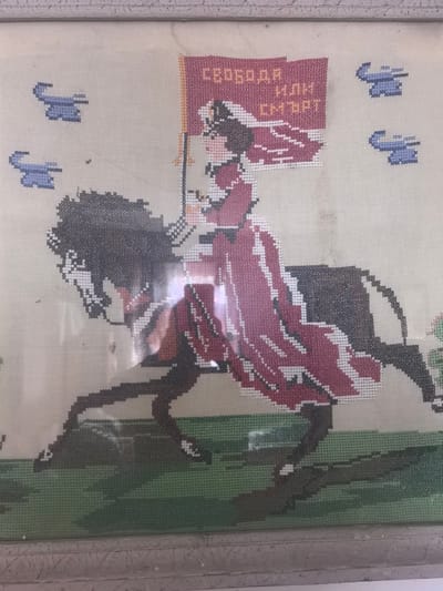 This is a framed cross-stitch embroidery depicting a woman on horseback holding a banner that reads "СВОБОДА ИЛИ СМЪРТ" (Liberty or Death) in Cyrillic. The woman is dressed in a red gown and hat, and the horse is black. In the background, there are four small blue shapes that appear to be stylized airplanes. The ground is depicted with green and brown cross-stitch. The embroidery is in the village of Tserovo, Bulgaria.