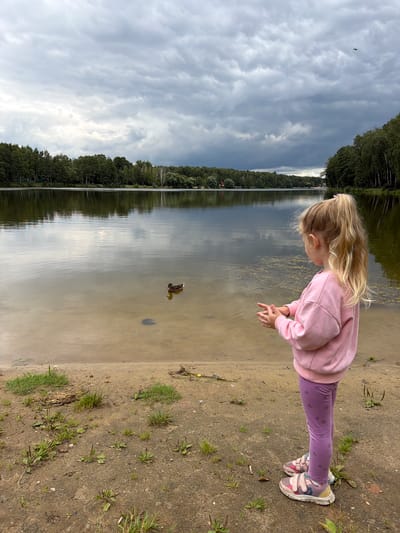 In Malakhovka, Russia, a young girl with blonde hair tied in a ponytail stands on a sandy shoreline, facing a lake. She wears a pink sweatshirt and purple leggings adorned with a bow pattern, and pink and white sneakers. Her hands are positioned in front of her as if she is about to toss something. In the lake, a duck swims near the shore. The water reflects the overcast sky and the dense green forest that lines the opposite bank. The foreground is a sandy bank with scattered patches of grass and some debris.