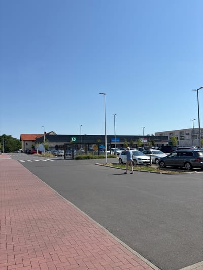 The image depicts a bright, sunny day in Elsenfeld, Germany, with a clear blue sky. The scene is set in a parking lot adjacent to a commercial building. A single person, a man wearing a blue t-shirt and beige pants, is the main subject, captured in motion as he appears to be walking or possibly dancing. He is facing away from the camera, looking towards the building.
The commercial building in the background features a dark facade with large windows and signage. One prominent sign displays a green rectangle with a white letter "D." Other visible text on the building includes "Mist-Lady" and various logos that are partially obscured. A row of parked cars lines the area in front of the building, and further back, trees and other buildings are visible.
The foreground is dominated by a paved area, transitioning from a dark asphalt road to a red brick sidewalk. Streetlights are positioned along the edge of the parking lot. The time of day is clearly daytime, suggested by the strong sunlight casting distinct shadows. The overall impression is one of a typical day in a suburban commercial area.