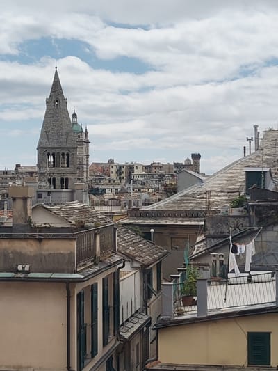 The image depicts an urban landscape in Genoa, Italy, characterized by a dense arrangement of buildings with varying heights and architectural styles. In the background, a prominent stone tower with a pointed spire rises above the surrounding structures. The foreground features rooftops of adjacent buildings with tile patterns, chimneys, and vegetation. Laundry hangs on a clothesline on one of the rooftops. The sky is overcast with clouds.