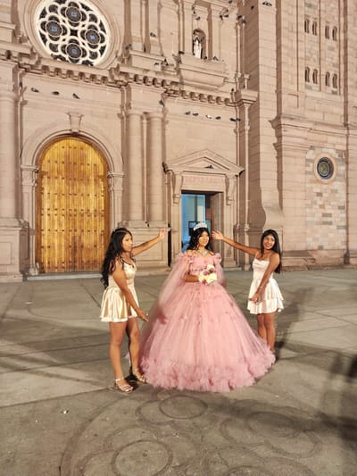 Three young women are posed for a photograph in front of the Cathedral of Tacna, located in Tacna, Peru. The central figure is a girl in a large, ornate, pink quinceañera-style dress, complete with a tiara and bouquet. Flanking her on either side are two young women wearing short, light-colored dresses and sandals, each with one arm outstretched towards the central figure. The cathedral is a grand, stone structure with a large wooden door, a rose window, and numerous architectural details. There are several birds perched on the cathedral's facade. The ground is paved and shows signs of wear, with circular patterns and tire tracks visible. The lighting suggests it is evening or night.