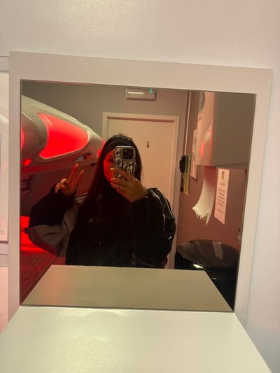 A mirror selfie captures an individual positioned in front of a white-framed mirror, which rests on a white countertop. The subject, likely female, wears a dark headband and a dark jacket. Their face is largely obscured by a smartphone with a reflective, patterned case, held up to take the photograph. The individual's left hand is raised, forming a peace sign (V-sign); the fingernails are painted yellow-orange.
Reflected in the mirror is an interior room, identified as a tanning salon booth in Temse, Belgium. Key features include a large, white-and-silver modern tanning capsule visible on the left, emitting a strong red light. In the background, a light-colored door is visible, above which a rectangular occupancy indicator displays a green light. To the right, wall-mounted fixtures include a white dispenser and a white paper towel unit with a torn sheet hanging, positioned above a dark, lidded waste bin. The room's walls are light-colored, predominantly illuminated by the red glow emanating from the tanning unit.