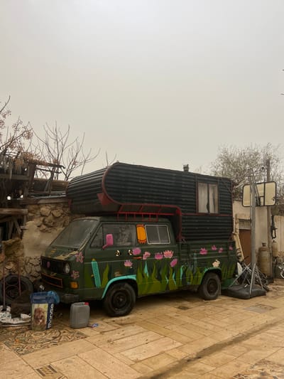 This image depicts a cluttered outdoor scene in Jisr az-Zarqa, Israel, under an overcast sky. The central subject is an olive green Volkswagen Transporter (T3 generation) pickup truck, extensively custom-modified into a camper vehicle. A large, dark-colored, corrugated metal camper unit with a curved front is mounted on the truck's bed, featuring a small window on its side and a chimney pipe on top. A red metal frame connects the camper unit to the truck's cab. The truck's body is decorated with colorful, folk-art style paintings including pink lotus flowers, green foliage, blue geometric shapes, yellow suns, and white rabbit motifs. Its side mirrors are pink.
To the immediate left of the truck's front wheel, a blue plastic trash bin featuring a dog's image and the word "monge" is visible. Two grey plastic jerry cans are on the ground beside it. Against a rough, stone wall to the left of the vehicle, stacked tires and other debris are present. Above the wall, tangled bare tree branches with some dried yellow fruits extend into the frame, along with wooden structures.
To the right of the vehicle, a freestanding basketball hoop with a white backboard and orange rim is positioned, with its dark base resting on the ground. Behind the hoop, various items including a tall grey gas cylinder, smaller gas cylinders, and metal scraps are visible. Further in the background, a light-colored, possibly dilapidated structure is seen, with sparse, leafless vegetation in the distance. The ground is paved with light brown, irregularly shaped tiles, showing signs of dampness or stains. No individuals are present.