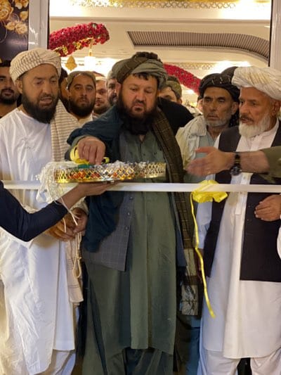 In قندهار, Afghanistan, a group of men are participating in what appears to be a ribbon-cutting ceremony. Most men are wearing traditional Afghan clothing, including turbans and long tunics. One man is pouring liquid from a small container onto a gold decorative tray held by an off-screen individual. A white ribbon with a yellow bow is stretched across the front of the scene. The background shows additional men and floral decorations. The setting appears to be indoors.