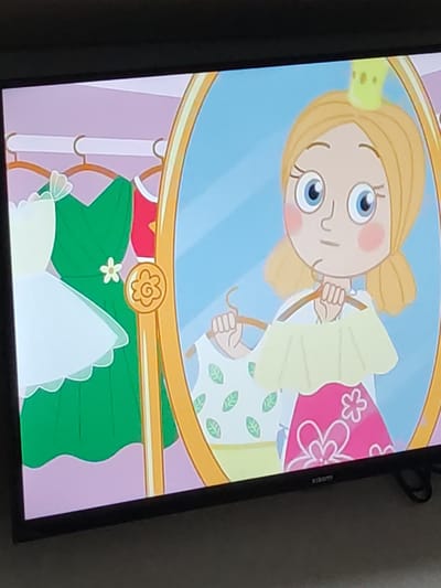 A cartoon image on a television screen depicts a young girl with blonde hair and blue eyes, wearing a small yellow crown. She is looking into a large, ornate mirror, holding a light yellow dress on a hanger. Her reflection in the mirror is identical. The background shows a closet with several dresses hanging on a rack. To the left of the mirror, a white and green dress is visible, and to the right, a pink dress with floral patterns. The overall scene appears to be a dressing room. There is no discernible location in the city of Tomsk, Russia, as the image is a digital illustration.