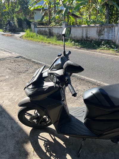 The image depicts a dark-colored scooter parked on a concrete surface next to a paved road. The scooter is in the foreground, with its front wheel and handlebars visible. A shadow of the scooter's wheel is cast on the ground. In the background, a road curves to the left, with a person on another motorbike visible in the distance. Lush green vegetation, including banana trees, and a low, weathered wall are situated along the side of the road. The scene appears to be outdoors in a tropical or subtropical environment, possibly in a rural or semi-rural area. Given the context of Pagowan, Indonesia, this is likely a street scene in or near the village.
