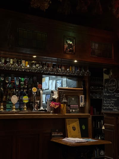 The image depicts the interior of a dimly lit pub or bar, located in Norton, United Kingdom. The scene is devoid of people. The environment is characterized by extensive dark wooden paneling across the walls and bar structure. Suspended from the ceiling, partially visible at the top, are dried botanical arrangements.

Above the main bar, two dark rectangular window frames and a smaller, illuminated stained-glass window are set into the wooden paneling. Beneath this, a row of empty wine glasses hangs upside down, positioned in front of shelves stocked with numerous bottles of spirits and liqueurs.

The wooden bar top features multiple beer taps, including those for "BREW HOP ON" and "BROADOAK PERRY." A small bouquet of pink and purple flowers rests in a vase on the bar. Adjacent to the flowers is a brass bell. A framed sign, likely an award, mentions "BEST BARN" and "BEST EVENTS TEAM." Behind the bar, a tiled wall displays various tacked papers and a small blackboard with the faint handwritten word "pork."

To the right, a freestanding blackboard titled "COCKTAIL DRINKS MENU" lists several cocktails, including "The Pegasus - Our Moscow Mule," "The Quince Essential," "The HH Garden," "A Whiskey Mac," and "Espresso Martini/After 8 Martini." Below this, shelves hold glassware and plates.

In the foreground, a wooden counter extends from the main bar, holding a stack of white papers and two menu-like folders, one yellow and one dark green. The overall lighting is dim, utilizing recessed spotlights above the bar. No specific actions or interactions are occurring.