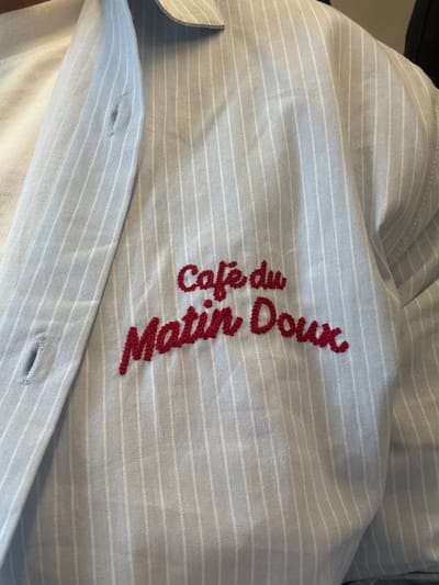 A close-up shot reveals a light blue and white striped shirt with the embroidered words "Cafe du Matin Doux" in red thread. The shirt's texture and stitching are visible, and the fabric appears slightly rumpled. A button and buttonhole are also visible along the shirt's placket. The image does not provide any information about the location in Peschiera Borromeo, Italy, nor does it depict any people, subjects, actions, or interactions. The environment is limited to the shirt itself.