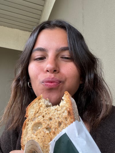 A young adult female is depicted in a medium close-up, holding a thick slice of toasted bread, partially consumed, wrapped in white paper with a green section. She has long, dark, wavy hair, dark eyes, and a light-to-medium complexion. She is wearing a dark brown garment and a silver-colored hoop earring in her left ear. Her lips are pursed, and her eyes are partially closed, conveying a distinct expression. The background features a light beige or off-white textured wall on the right and a slatted ceiling structure on the upper left, suggesting an indoor or covered outdoor area. The specific location in the city of West Windsor, United States, is not discernible from the visual information provided within the image.