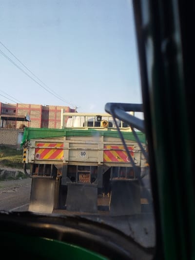 The image displays the rear view of a truck observed from the interior of another vehicle, situated on a dirt road in Keekonyokie ward, Kenya. The truck is predominantly off-white with a green tarp covering its cargo bed. Its rear features yellow and red diagonal reflective striping, a white square sign indicating "80 KPH," and a Kenyan license plate numbered "KDA 092G." Two circular red reflectors are visible, and large black mud flaps hang behind the rear wheels. In the background, under a clear blue sky, are several buildings, including a multi-story structure with exposed light-colored block walls patterned with red accents, suggestive of ongoing construction or unpainted finishes. Utility lines are visible horizontally across the upper portion of the background. Sparse vegetation grows on the ground near the buildings. No individuals or specific actions are discernible.