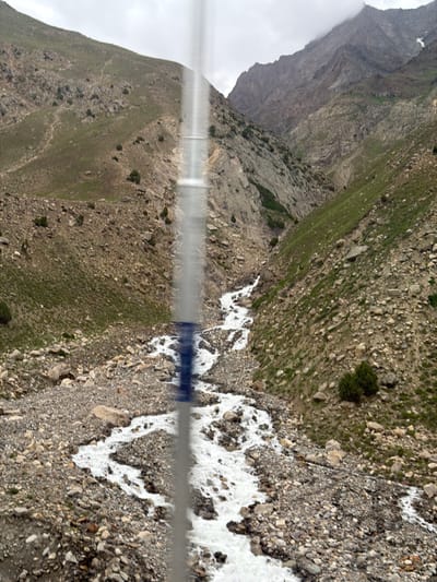 The image depicts a mountain landscape with a river flowing through a rocky, sparsely vegetated terrain. A vertical, blurry element, possibly a pole or a streak of light, runs through the center of the image, obscuring parts of the scene. The mountains are steep and rugged, with patches of green grass and scattered rocks. The river is white and turbulent, indicating a fast current. The sky is overcast, suggesting cloudy weather. The image is likely taken in a rural or mountainous region, given the natural landscape and absence of urban elements. The location is identified as Chhika-Be, India, which is a mountainous area known for its natural beauty and rivers.
