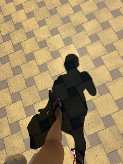 The image shows a person's shadow and legs walking on a yellow and gray brick pathway. The person is wearing flip-flops and is carrying a bag. The scene appears to be outdoors on a sunny day. The location is the city of Timashevsk, Russia.
