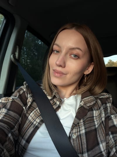 A woman is in a car, wearing a brown and white plaid shirt over a white t-shirt, and a seatbelt. She has light brown hair, fair skin, and green eyes. Her left ear has a gold chain-link earring. The background shows a window with greenery outside. There is no indication of the location being Rogachovo, Russia.