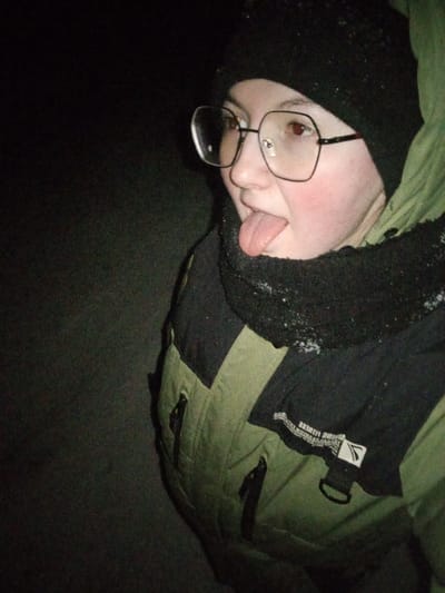 The image presents a close-up, eye-level portrait of an individual in a dark environment. Visual information to confirm the specified location of Старый Шагирт, Russia, is absent.
The subject is a young person with fair skin and flushed cheeks, wearing a black knit beanie, large clear-framed eyeglasses with dark temples, and an olive-green puffer jacket with black shoulder panels. The jacket features zippered pockets and a partially visible white text label on the chest. A black knitted neck warmer or high collar, speckled with small white particles consistent with snowflakes, is worn. The subject's mouth is open, with the tongue extended. Both eyes exhibit a red-eye effect, indicating flash usage in low light. The background is uniformly dark and devoid of features.