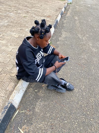 A young Black person, with their hair styled in multiple Bantu knots, sits on a textured concrete curb next to a paved asphalt surface. They are wearing a black long-sleeved shirt with white stripes on the sleeves and a white trim around the neckline, and dark-wash jeans. The individual is holding a blue smartphone in their right hand, looking down at the screen. On their left wrist, a beaded bracelet with a cross charm is visible. They are wearing black, glossy loafers with silver buckles and thick, lug soles. The background shows a continuation of the paved surface and a black and white striped curb. The scene appears to be outdoors, possibly on a street or public walkway in Zawan, Nigeria.