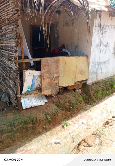 In Takau, Nigeria, a rustic dwelling is depicted. The structure features a roof made of dried palm fronds and walls constructed from bamboo and mud. In front of the dwelling, a discarded television box is visible. The ground is a mix of soil and moss, with scattered debris. The overall scene conveys a sense of poverty and dilapidation.