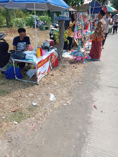 The scene is a street market in Kendal, Indonesia. There is a food stall with a young man seated behind it. The man is wearing a grey shirt and black pants. He has short black hair. He is looking towards the viewer. The stall is made of a folding table with a white tablecloth. It is decorated with a banner that says "Sosis Tel Kombo" in red, white, and green lettering. There are red and yellow condiments in front of the man. Behind the food stall, there is a large tree with a blue umbrella. A woman is standing next to the tree. She is wearing a black and white long-sleeved shirt. She has long black hair. She is looking down at the viewer. She is holding a white plastic bag. A small motorcycle is parked behind her. In the background, there is a group of people. They are standing in front of a street vendor selling miscellaneous items from a table. The street vendor is wearing a brown long-sleeved shirt. He is looking to the left. He has a shaved head. He is wearing a blue and white sarong. The vendor is selling various items including toys, clothes, and food. The street is lined with concrete slabs and is covered in dry leaves. There are a few plastic bags on the ground. The sky is clear and blue. The location is in the city of Kendal, Indonesia. The coordinates are -6.934 latitude and 110.203 longitude.