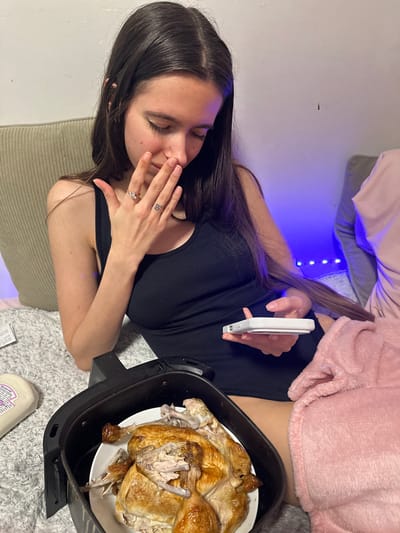 The scene depicts an indoor residential setting in Corby, United Kingdom. A young female subject, positioned centrally, is seated on a textured gray blanket. She has long, dark brown hair and is wearing a black tank top. Her right hand is covering her mouth and nose, and her eyes are cast downwards. A ring with a clear stone is observed on her left ring finger, and a small, dark, stylized "ß" tattoo is visible on her right middle finger. Her left hand holds a white smartphone, which she is operating with her thumb. A pink fuzzy blanket covers her lower body.
In the immediate foreground, a black air fryer basket contains a white plate holding a roasted chicken that appears partially consumed or carved. To the left of the air fryer, a white plastic squeeze bottle with a partially visible purple label rests on the gray blanket. The background consists of a light-colored wall illuminated by a string of blue LED lights running horizontally along its lower section. A light beige upholstered cushion is visible behind the subject to the left, and a gray upholstered surface is partially visible to the right.
