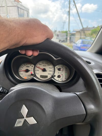 The image captures a close-up, first-person perspective from inside a vehicle, with a hand gripping the steering wheel. The focus is on the dashboard and steering wheel, with the road and scenery visible through the windshield.
The steering wheel is dark gray and appears to be made of plastic or leather. The Mitsubishi logo is prominently displayed in the center of the steering wheel. To the left of the steering wheel, a windshield wiper stalk is visible.
The dashboard features two prominent gauges. The left gauge displays an RPM meter with a red needle pointing to the left, indicating a low engine speed. Several indicator lights are visible below the RPM meter, with one orange light illuminated. The right gauge is a speedometer, with a red needle indicating a speed of approximately 40 km/h. Below the speedometer, a digital display shows the time as 03:25:03.
Through the windshield, a blurred view of a street scene is visible. A blue car is parked in the background. A telephone pole and wires are also visible. The sky is partly cloudy. The overall lighting suggests it is daytime. The location is identified as Sergey-Pole, Russia.