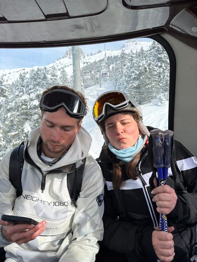Two individuals are seated inside a ski gondola cabin, traversing a snow-covered mountainous region in a ski resort near Les Allues, France. The male, positioned on the left, wears a white and grey patterned hooded jacket with "NEIGHTY 1080® GEAR" branding and black-framed ski goggles resting on his forehead. He is observed looking downwards at a black smartphone held in his left hand. A black backpack is visible on his shoulders. The female, positioned on the right, wears a black and white ski jacket, a light blue neck gaiter, and a grey ski helmet with orange-tinted goggles pushed up onto the helmet. She holds two blue and black ski poles, branded "IPRON" and "Ski-M.com", in her left hand and is making a puckered lip expression towards the viewer. Both individuals appear to be young adults. Through the gondola's window, the background reveals an extensive snow-covered landscape with dense coniferous trees and mountain slopes, under a clear sky, with a functional chairlift system visible in the distance.