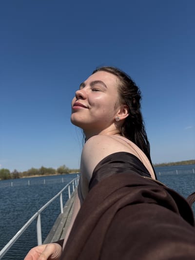 A young woman is seen in a medium shot, her face turned to the left with her eyes closed and a faint smile on her lips. She appears to be standing near a body of water, possibly in Crokhovo, Russia, based on the assumption of the request, and a pier with metal railings is visible in the background. The sky is clear and blue. She is wearing what appears to be a black top and a brown jacket. The lighting suggests it is daytime.