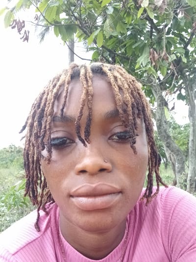 An overhead portrait-style shot captures a person with medium-brown skin and dreadlocks looking directly at the camera. The person's head is tilted slightly, and their expression is neutral. They have dark eyes and full lips. A small, clear or silver stud piercing is visible on the left side of their nose. Their dreadlocks are a mix of dark and lighter brown hues, with some strands falling across the forehead and around the face.
The person is wearing a pink, ribbed top, which features a horizontal striped texture. A thin gold chain is visible around their neck, resting on the fabric of the shirt. The background consists of lush green foliage and tree branches, indicating an outdoor setting, possibly in a garden or natural area. The sky in the upper right portion of the image is bright and appears overcast or hazy white. The lighting suggests it is daytime. The image hints at a location like Oba-Ile, Nigeria, given the natural outdoor environment. No visible text is present in the image.