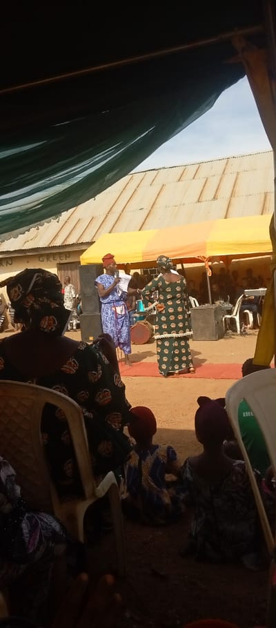 The image depicts an outdoor event in Kasuwan Magani, Nigeria. The scene is observed from under a dark green tarp, which partially shades the foreground. An audience is present, with individuals seated on white plastic chairs or kneeling on the dirt ground; an adult female in a dark green patterned dress and headwrap is prominently visible from behind in the left foreground.
In the midground, a makeshift stage is set on a red mat over dirt, under a yellow and orange striped canopy tent. Two central figures stand on the stage. On the left, a male in a blue patterned outfit and a reddish-orange headwrap holds a microphone, facing right. To his right, a female in a dark green patterned dress with white trim and a headwrap stands, facing the male. Two large black speakers flank the stage, with a drum and a keyboard visible among other equipment and seated individuals further back under the canopy.
The background features a single-story building with a light-colored wall and a corrugated metal roof. Green text "RN GREEN" is visible on the building wall. The sky above is clear with scattered clouds.