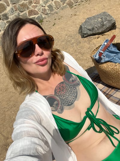 In this overhead shot, a person lies on sandy ground near a cobblestone path, bathed in bright sunlight suggesting a warm, clear day. The person wears a green bikini top and a sheer white cover-up. They have on large, brown-tinted sunglasses and their makeup includes a touch of pink lipstick. A prominent tattoo adorns their chest, featuring a complex geometric design with circular elements and red accents.

To the upper left of the frame, a woven straw bag is visible, containing folded denim jeans and a brown leather belt. A large, rough-textured gray rock rests on the sand near the bag. A checkered blanket or towel is partially visible behind the person.

The scene suggests a leisurely day outdoors, possibly at a beach or park, given the natural elements and casual attire. The lighting indicates it is likely midday. There is no visible text in the image. The setting appears to be outdoors in a sunny environment.