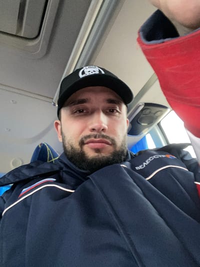 A man with a beard, wearing a black cap with a white logo, a navy blue jacket with a white stripe and red, white, and blue details, is in what appears to be the interior of an airplane. The man is looking towards the camera. The interior walls of the aircraft are visible. The location in Koskolovo, Russia is not determinable from the image.