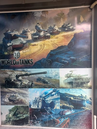 This is a poster featuring various images of tanks, predominantly from the video game "World of Tanks." The poster is divided into a grid of images, with some larger and some smaller.
The dominant imagery on the left side of the poster depicts a line of tanks advancing through a tropical or desert-like environment with palm trees and sand. The lighting suggests it could be daytime, possibly with dust or smoke in the air, creating a sense of action and intensity. The tanks appear to be in motion, with one in the foreground prominently displaying its barrel and tracks.
The right side of the poster is a collage of smaller images, also featuring tanks in different settings. Some show tanks in more urban or rocky terrains, while others are close-ups of tank components or action shots. The overall mood conveyed by the poster is one of power, conflict, and possibly excitement, aligning with the theme of a war-themed video game.
The poster displays text in English. Prominently featured in large, stylized lettering is "WORLD OF TANKS." The "World of Tanks" logo, a silver shield with a stylized "W" inside, is also visible.
Given the location context of Kasansay, Uzbekistan, this poster is likely displayed in a public space, perhaps a shop or entertainment venue, catering to fans of the game or military vehicles.