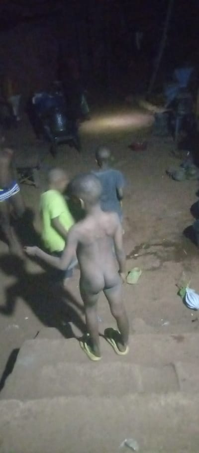 In a poorly lit outdoor setting, several children are present. One child is completely nude from the waist down, wearing only shoes. Another child is wearing a lime green shirt. A third child has on a dark blue or black shirt. The ground appears to be dirt or sand. In the background, there are objects that look like chairs or equipment, partially obscured by the darkness.