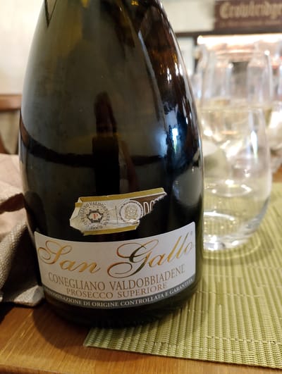A close-up, eye-level, slightly angled shot shows a dark green glass wine bottle standing upright on a wooden table covered with a light green woven placemat. The bottle is the primary subject, positioned in the foreground and taking up most of the frame. A label on the bottle has gold cursive lettering that reads "San Gallo", followed by "CONEGLIANO VALDOBBIADENE PROSECCO SUPERIORE". A smaller, rectangular seal with official markings is affixed to the upper portion of the label. Two clear wine glasses, one behind the other, are visible in the background on the right, each containing a small amount of clear liquid. The lighting is warm and ambient, creating reflections on the bottle's surface. In the far background, a blurry sign with cursive text is visible. The scene appears to be indoors, possibly at a restaurant or winery, with a casual, relaxed atmosphere. The specific location in the city of Torri in Sabina, Italy, is not discernible from the visual information provided in the image.