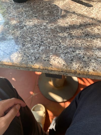 The image displays a close-up, eye-level perspective from a seated individual. A speckled granite tabletop, predominantly grey and reddish-brown with dark granular inclusions, occupies the upper portion of the frame, exhibiting direct sunlight and distinct shadows. The table's front edge is rounded and shows signs of wear. Beneath the table, a dark, cylindrical metal pedestal table base with a circular foot is visible on a reddish-brown tiled floor, which also displays sharp sunlit areas and shadows. In the lower-left, a person's left hand, with fair skin, rests on their dark-colored upper thigh, clad in dark fabric pants. To the lower-right, a light-colored, possibly beige or off-white, slip-on sandal is present on the tiled floor. No specific details or landmarks in the image allow for identification of a precise location within the city of Đak Đoa, Vietnam.