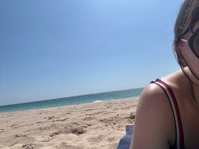 The image displays a beach scene in Phra Ae, Thailand, under a clear, bright blue sky. The foreground, positioned in the lower right, features the partial view of a person's left shoulder and the side of their head. The person wears dark-rimmed sunglasses and a swimsuit top with a red, white, and black striped strap over their shoulder. A corner of a dark blue towel or mat is visible beneath them on the sand. The midground consists of light brown sand, extending towards the water. In the background, the turquoise-blue ocean stretches horizontally, with small waves visible at the shore. Several indistinct figures, likely people, are visible in the water. The upper half of the image is dominated by a cloudless, light blue sky.