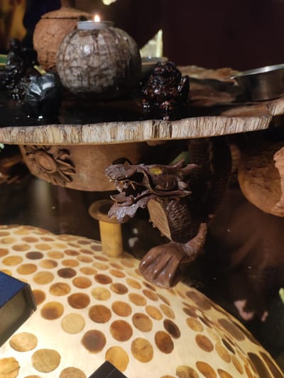 A wooden sculpture of a dragon with green eyes is positioned on a wooden table under an ornate wooden platform. The platform holds several decorative items including a light-colored, round candle holder with a lit candle, a dark round object, and an urn-like item. The dragon sculpture rests on a small, cylindrical wooden stand. Underneath the dragon is a lamp with a circular pattern of brown spots on a light background. The location in the city of Lokaserena, Indonesia cannot be determined from the image.