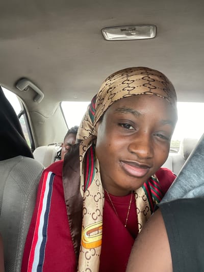 The media file shows a close-up of a young Black woman inside a vehicle. She is wearing a headscarf with a repeating logo pattern and a red long-sleeved shirt with striped trim. The woman is smiling and looking towards the camera. The interior of the vehicle is visible, including a gray ceiling with a light fixture and a grab handle. Another person is partially visible in the background, also inside the vehicle. The lighting suggests it is daytime, possibly overcast. The overall scene appears to be a casual moment within a car, likely in Kwali, Nigeria, as indicated by the provided location context. There is no visible text in the image.