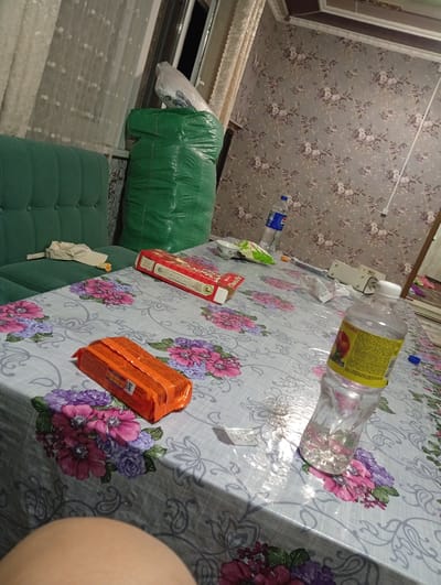 The image depicts an indoor scene, likely a domestic setting, with a table covered by a floral tablecloth. On the table are several items, including a red box, an orange package, and two plastic bottles of liquid, one of which is partially filled and has a yellow label. There are also some scattered wrappers and a small white cup. In the background, a green sofa is partially visible, and a large green plastic bag is standing next to it. The wall is covered with floral wallpaper. The lighting suggests it is either evening or nighttime. The image is taken from a low angle, showing a person's leg in the foreground. There is no specific indication of the city of Karavan, Kyrgyzstan.