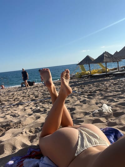 This image depicts a beach scene in Zankovići, Montenegro. In the foreground, a person lies on the sand, with their legs crossed and raised. They are wearing a light-colored bikini bottom. The sand is textured with ripples and footprints. In the mid-ground, there are several thatched umbrellas providing shade, and yellow pedal boats are visible behind them. The sea is calm and blue, meeting a clear sky. In the background, a man in a black t-shirt and shorts walks along the shoreline. Other people and beach structures are also present in the distance. The overall scene conveys a relaxed atmosphere on a sunny day at the beach.