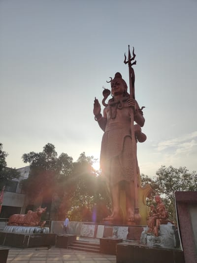 The image captures a low-angle, wide shot of the colossal statue of Lord Shiva at the Shiva Temple in Sector 29, Gurgaon, India. The sun is setting, casting a warm glow and lens flares across the scene, with trees and a building visible in the background. The statue, made of reddish-brown material, depicts Lord Shiva in a standing pose, holding a trident and a damaru, with snakes around his neck and arm. To the left of the main statue, a smaller statue of a bull is also visible. To the right, at the base of the main statue, there are smaller sculptures of deities seated on rocks. A person in a light-colored shirt walks in the middle ground, appearing to observe the statues. The ground in the foreground is paved.
