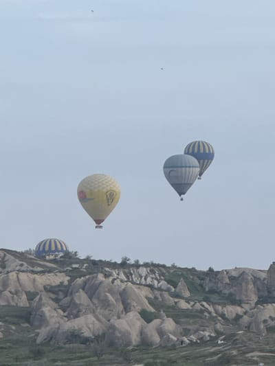 The image captures the Cappadocia region in Göreme, Turkey, with hot air balloons in the sky near the geological formations. The sky is overcast, the balloons are striped and colored, and the rocks are textured and eroded.