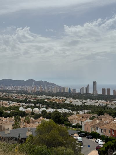 This is a daytime, high-angle, wide shot of the city of Urbanització Golf Badia, Spain. The sky is overcast with white and grey clouds. In the distance, a range of dark, undulating mountains forms the horizon. Beyond the mountains, the sea is visible, appearing as a pale grey expanse.
The middle ground of the image is dominated by a densely packed cityscape featuring numerous high-rise buildings, many of which are residential towers. The buildings vary in height and architectural style, with some appearing modern and sleek, while others have a more traditional design. Some of these buildings are located along the coastline.
In the foreground, the scene transitions to a residential neighborhood characterized by smaller, terracotta-roofed houses with light-colored facades. These houses are surrounded by lush green trees and manicured hedges. A paved road curves through the neighborhood, with several cars parked along its edge, including a black SUV, a black sedan, a white sedan, and a white SUV. A street sign with a blue and white symbol is visible on the side of the road. The foreground also shows areas of dry grass and other vegetation, suggesting a hilly terrain. The overall impression is of a sprawling urban and suburban environment set against a natural mountainous backdrop.